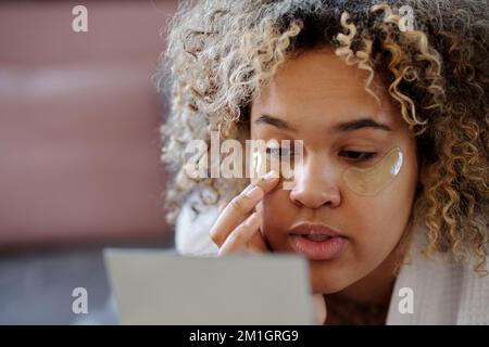 Gros plan de la jeune femme en train de s'hydrater sous les paupières sur le visage tout en se relaxant sur le lit après l'hygiène du matin et en regardant dans le miroir Banque D'Images