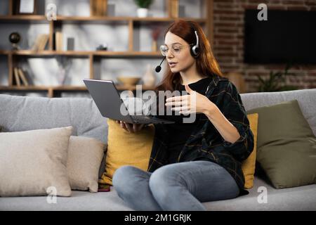 Vue de face d'une fille heureuse de cheveux rouges dans le casque étudier avec un ordinateur portable à la maison, parler tout en regardant l'appareil photo avec un entraîneur ou un professeur de cours d'apprentissage à distance. Éducation en ligne pour les femmes de race blanche. Banque D'Images