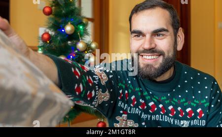 Heureux barbu homme surprenant avec un cadeau enveloppé devant un arbre de noël portant un chandail de noël Banque D'Images