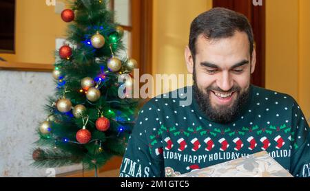 Heureux barbu homme surprenant avec un cadeau enveloppé devant un arbre de noël portant un chandail de noël Banque D'Images