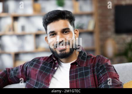 Gros plan portrait d'un beau barbu assis, reposant sur un canapé confortable dans un salon moderne. Jeune homme du Moyen-Orient souriant, locataire ou locataire, détendez-vous sur le canapé à la maison, profitez du week-end. Banque D'Images