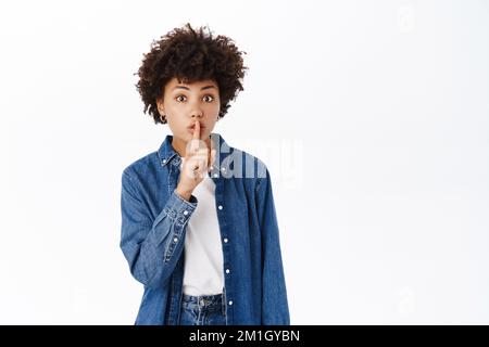 Portrait de la femme noire montre hush, shhh shush geste, tabou, demande à garder calme, gossiping, se tient sur fond blanc Banque D'Images