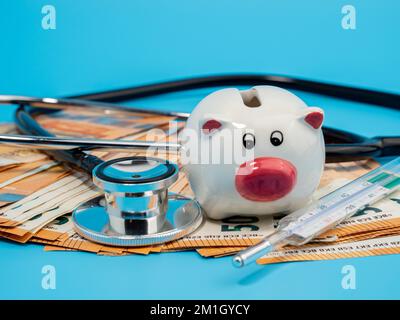 Stéthoscope médical, banque de porc, thermomètre et billets sur fond bleu. Gros plan. Banque D'Images