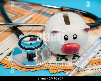 Stéthoscope médical, banque de porc, thermomètre et billets sur fond bleu. Gros plan. Banque D'Images