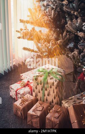 Les cadeaux enveloppés sous un arbre de Noël tandis que les rayons du soleil brillent à travers les stores verticaux sur les portes de patio à côté de l'arbre. Banque D'Images