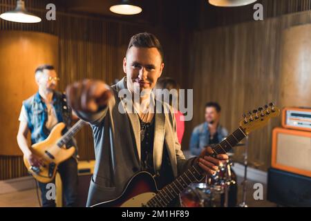 Guitariste mâle caucasien positif en blouson gris tenant sa guitare électrique et pointant vers la caméra avec l'index. Main floue au premier plan. Bande de garage. Photo de haute qualité Banque D'Images