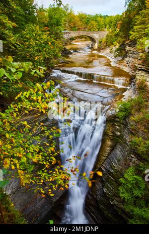 Cascade dans des gorges profondes avec feuillage d'automne, forêt, et pont voûté en pierre en arrière-plan Banque D'Images