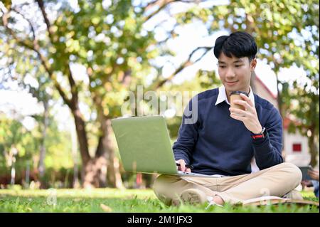 Jeune asiatique beau et intelligent étudiant à l'université en utilisant un ordinateur portable, en sirotant un café, en faisant des devoirs tout en étant assis dans le parc du campus. Banque D'Images