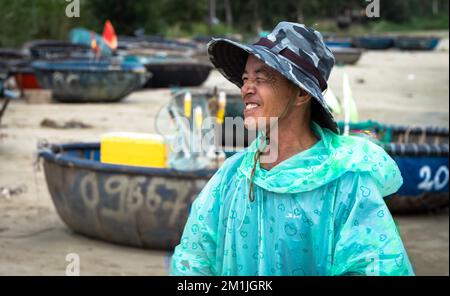 Un pêcheur vietnamien portant un imperméable avec des coracles traditionnels utilisés pour pêcher un jour de pluie sur la plage My Khe à Danang, Vietnam. Banque D'Images