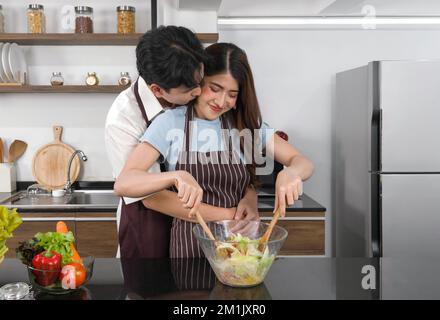 Un couple asiatique passe du temps ensemble dans la cuisine. Le jeune homme embrasse et embrasse la joue de sa petite amie de derrière tandis que la femme mélange les légumes dans un Banque D'Images