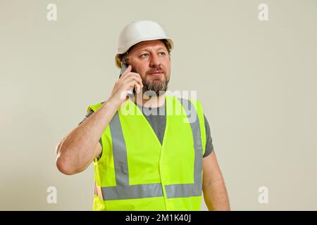 Homme travailleur dans un gilet et un casque de construction blanc qui parle au téléphone sur un fond blanc Banque D'Images