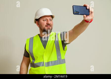 Un homme travailleur dans un gilet et un casque de construction blanc prend un selfie sur le téléphone sur un fond blanc Banque D'Images