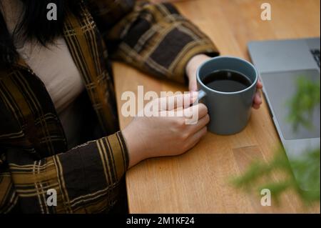 Coupe courte, Une femme asiatique taille basse en chemise en flanelle tenant une tasse de café, assise à son bureau. Banque D'Images