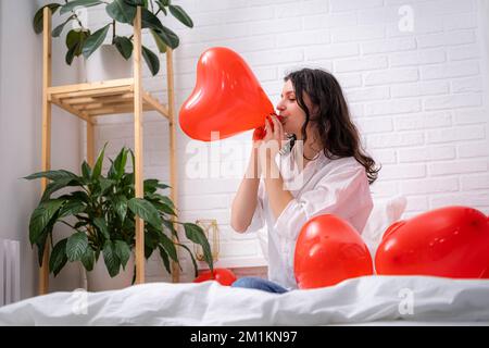 Fille est souffle air ballon à la maison beau brunette fille gonflant rouge coeur en forme de ballon assis sur le lit. Préparation pour la fête de la Saint-Valentin Banque D'Images