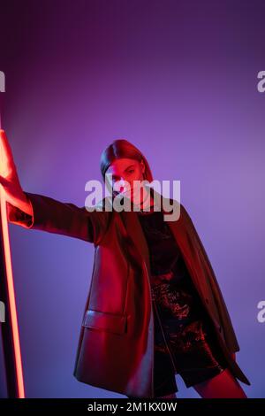 jeune femme en mini jupe et veste de cuir debout près de la lampe néon vibrante sur fond violet, image de stock Banque D'Images