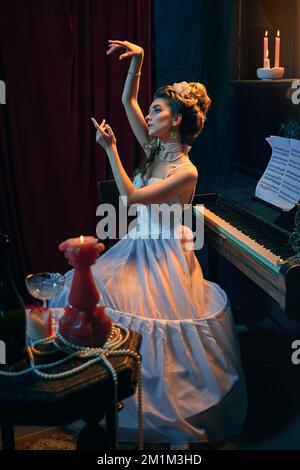 Portrait de la belle jeune femme à l'image de la personne médiévale en robe blanche tendre assise au piano. Style de vie de princesse. Comparaison d'époques, beauté Banque D'Images