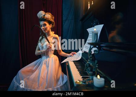 Portrait de la belle jeune femme à l'image de la personne médiévale en robe blanche tendre assise au piano avec des notes et des quill. Faire une jolie mélodie Banque D'Images