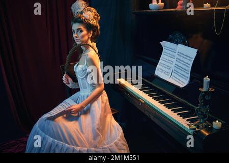 Portrait d'une jeune femme élégante à l'image d'une personne royale médiévale posant dans une robe blanche tendre, assise au piano dans une pièce de style vintage. Comparaison Banque D'Images