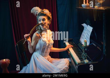 Réunion en soirée. Portrait de la belle jeune femme à l'image de la personne médiévale en robe blanche tendre assise au piano et jouant. Comparaison des époques Banque D'Images