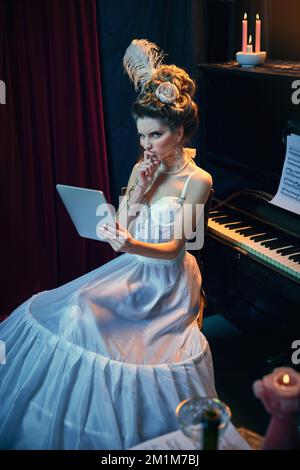 Lecture de romans. Portrait de la jeune belle femme en mage de la personne médiévale en élégante robe blanche, assise au piano et posant avec un comprimé Banque D'Images