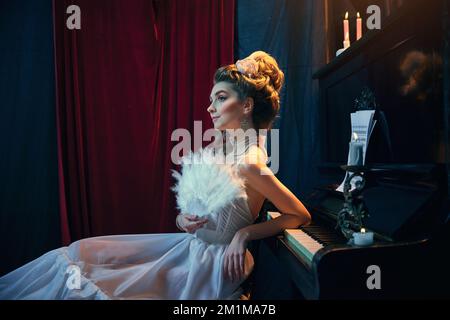 Femme tendre. Portrait de la jeune belle fille à l'image de la personne médiévale en élégante robe blanche assise au piano. Comparaison d'époques, beauté Banque D'Images