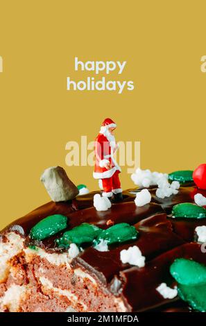 le texte happy holidays et un père noël miniature debout à côté d'un sac plein de cadeaux, sur un gâteau de bûche de yule devant un fond jaune Banque D'Images