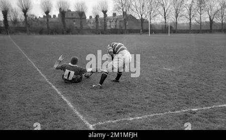 1950s, historique, un joueur de rugby en maillot de basket-ball blindé le ballon par la ligne de contact, après avoir retiré son adversaire, un avant-poste avec le numéro un sur son maillot qui est sur le terrain, dans un match entre deux clubs amateurs, Wasps FC v Wimbledon RFC, Londres, Angleterre, Royaume-Uni. Wimbledon Hornets - comme on l'appelait alors - étaient l'un des 21 membres originaux de l'Union de football de Rugby (RFU) fondée en 1871. Bien que fondée quelques années plus tôt en 1867, Wasps football Club (Wasps FC) pour des raisons pas entièrement claires, n'est pas devenu un membre fondateur de la RFU mais est devenu membre en 1872. Banque D'Images