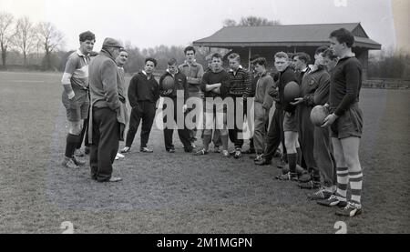 1950s, historique, membres de l'équipe de l'Union de football de rugby des écoles anglaises lors d'une session d'entraînement à l'extérieur sur un terrain debout ensemble obtenant des instructions de l'un des deux entraîneurs masculins adultes présents, Angleterre, Royaume-Uni. Tous les garçons sont dans le groupe plus de 15, avec certains en short, mais beaucoup portent les survêtements de coton bougé de l'époque. Banque D'Images
