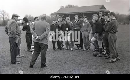 1950s, historique, membres de l'équipe de l'Union de football de rugby des écoles anglaises lors d'une session d'entraînement à l'extérieur sur un terrain debout ensemble obtenant des instructions de l'un des deux entraîneurs masculins adultes présents, Angleterre, Royaume-Uni. Tous les garçons sont dans le groupe plus de 15, avec certains en short, mais beaucoup portent les survêtements de coton bougé de l'époque. Banque D'Images