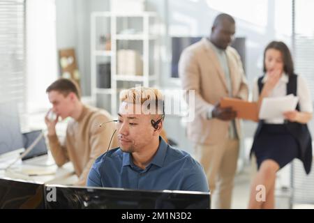 Grand angle de vue jeune homme asiatique portant un micro-casque au bureau et regardant l'écran d'un ordinateur tout en travaillant dans le service clientèle du centre de surveillance Banque D'Images