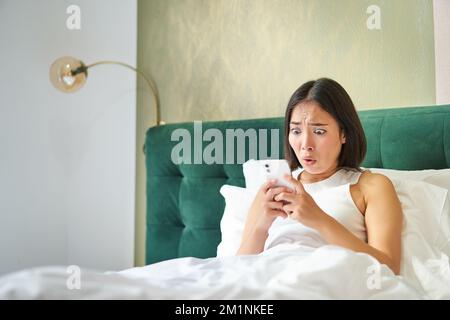 Portrait d'une femme coréenne choquée regardant son téléphone mobile, couché au lit, lisant de mauvaises nouvelles choquantes sur l'application pour smartphone Banque D'Images