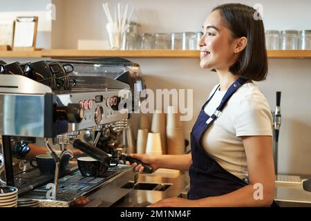 Bonne fille asiatique, le barista en tablier travaille avec une machine à café derrière le comptoir, debout dans un café et prépare du cappuccino Banque D'Images