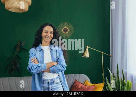 Jeune belle femme hispanique à la maison souriant et regardant la caméra avec les bras croisés, portrait de femme avec les cheveux bouclés dans le salon. Banque D'Images