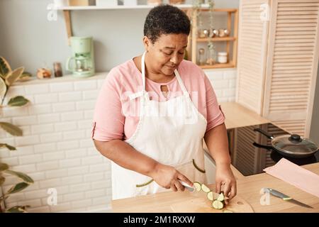 Portrait en grand angle de femme sénior noire coupant des légumes dans une cuisine confortable, tons chauds Banque D'Images