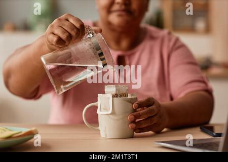 Gros plan de la femme noire senior faisant le thé dans la cuisine maison confortable, tenant la bouilloire en verre et l'eau de verser, l'espace de copie Banque D'Images