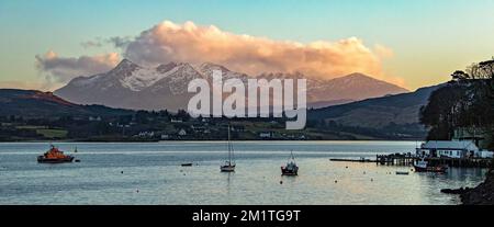 Vue sur le port de Portree vers Camustianavaig avec de petits bateaux en premier plan, des maisons périphériques et la lumière du soleil orange sur les nuages et Ben Ti Banque D'Images