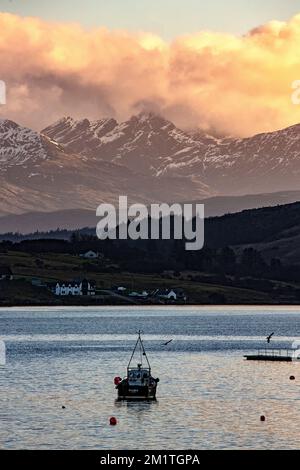 Vue sur le port de Portree vers Camustianavaig avec un petit bateau de pêche en premier plan, des maisons périphériques et la lumière du soleil orange sur les nuages A. Banque D'Images