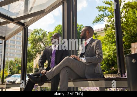 Deux hommes d'affaires à la peau sombre en costume communiquent. Les amis et les partenaires d'affaires sont assis sur un banc dans la ville sur fond de Banque D'Images