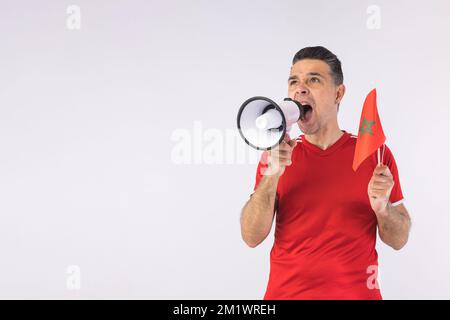 Homme portant un t-shirt rouge, criant dans un mégaphone et tenant un drapeau marocain. Sport, coupe du monde et concept de fan. Banque D'Images