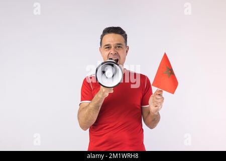 Homme portant un t-shirt rouge, criant dans un mégaphone et tenant un drapeau marocain. Sport, coupe du monde et concept de fan. Banque D'Images