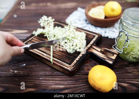 La main d'un herboriste coupe les ingrédients pour faire une boisson rafraîchissante à partir de fleurs de sureau. Boisson d'été à base de fleurs fraîches. Banque D'Images