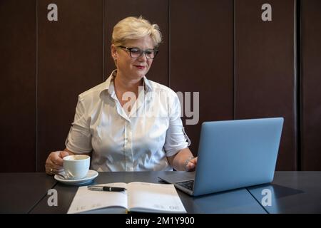 Femme caucasienne d'âge moyen avec des cheveux courts et des lunettes travaillant devant un ordinateur portable et une travailleuse professionnelle souriante et à distance appréciant une tasse de café Banque D'Images