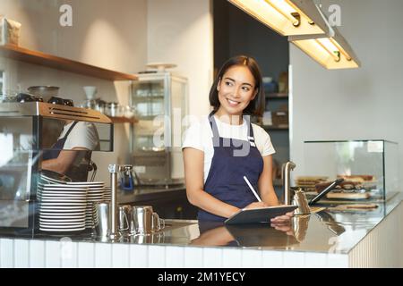 Portrait d'une belle fille asiatique souriante, barista dans un café travaillant derrière le comptoir, utilisant une tablette comme terminal de point de vente, traitement de l'ordre Banque D'Images