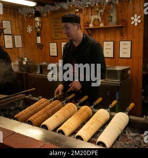 Budapest, Hongrie - 11 novembre 2022: Un jeune homme qui cuit des gâteaux de cheminée hongrois sur des charbons chauds dans un marché de noël à Budapest. Banque D'Images