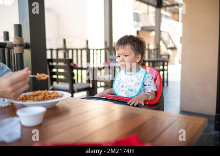 Tout-petit assis sur une chaise haute dans le restaurant pendant un repas. Beau garçon multiracial de un an et demi ayant un repas à l'extérieur Banque D'Images