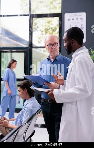 Un médecin afro-américain discutant de l'expertise médicale d'un patient senior lors de la consultation de visite de contrôle à l'hôpital. Medic expliquant le traitement des soins de santé pendant que l'homme signe des papiers d'assurance Banque D'Images