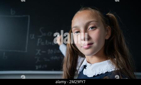 Portrait d'une fille de race blanche en classe. L'écolière écrit la formule avec de la craie sur le tableau noir et regarde dans l'appareil photo. Banque D'Images