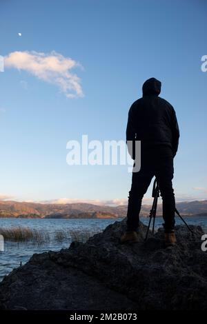 Une silhouette jeune homme photographe debout au-dessus d'un rocher avec un lac bleu, des montagnes et un croissant de lune cirant dans le coin supérieur gauche Banque D'Images