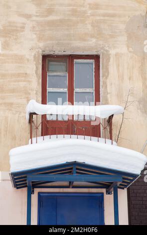 Un petit balcon couvert de neige sur une verrière avec une grande dérive de neige blanche. Ancienne porte vitrée en bois, balcon au-dessus de l'entrée. Hiver jour nuageux, lumière douce. Banque D'Images