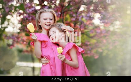 Les enfants heureux mangent de la crème glacée. Portrait de deux sœurs jumelles qui embrasent et rient sur fond d'arbre rose de fleur. Vacances d'été pour les enfants Banque D'Images